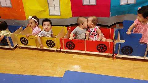 "A Baby Boy and Baby Girl Sit in A Toy Train and Kiss"