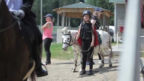 Little beautiful girl in a black suit and helmet stands near the pony and smiles