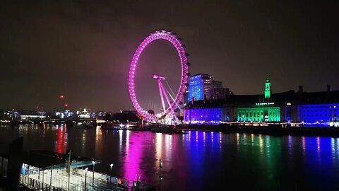 A rainy English night at the London eye, with a colourful light display in the background