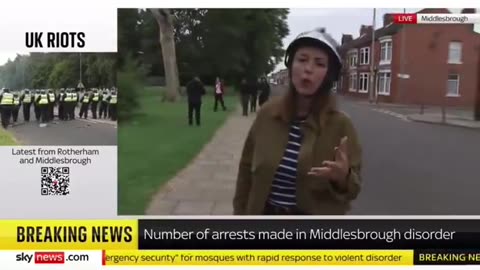 Armed migrants walk behind a reporter as she discusses "far-right" protestors