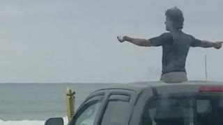 Guy does weird arm warmup while standing out sunroof of truck in front beach