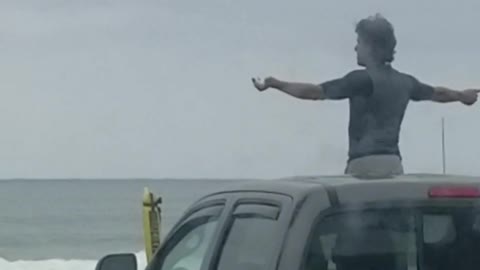 Guy does weird arm warmup while standing out sunroof of truck in front beach