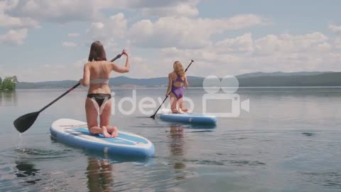 Rear View Of Two Girls In Swimsuit Doing Paddle Surfing On Knees In The Sea