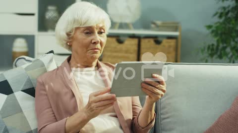 Senior Grey Haired Woman Having A Videochat On The Tablet Device Via Webcam In The Nice Living Room