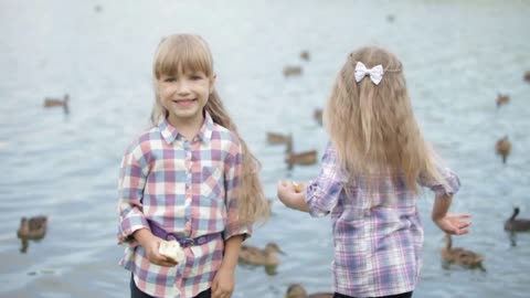 Two little girls feeding ducks at park turning around and smiling at camera