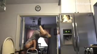 Guy tries to lift himself and balance between two chairs in kitchen but flips and hits wall