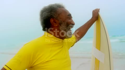 Front view of happy active senior African American man with surfboard standing on the beach