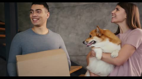 Smiling young man with box and woman with fluffy corgi dog in arms come into new spacious house
