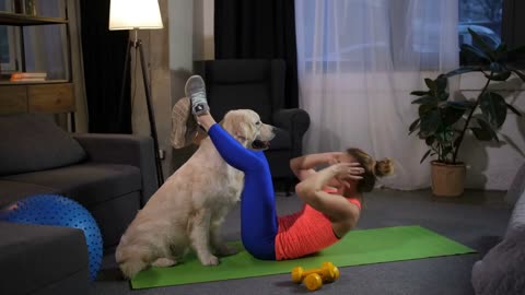 Doing fitness female lying on training mat keeping feet on labrador dog's neck in domestic room