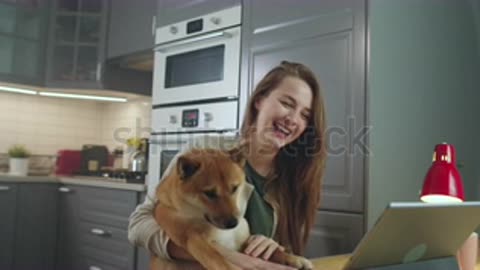 Side View of Young Woman Sitting at Table and Petting Her Dog While Making Video Call.