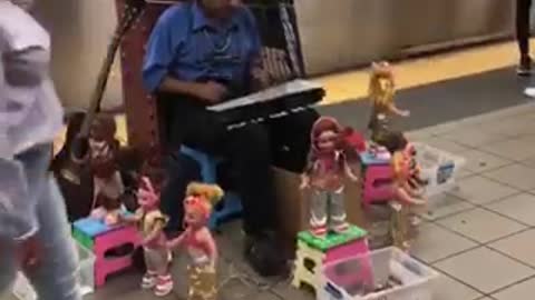 Guy blue shirt playing xylophone with a bunch of dancing dolls around him