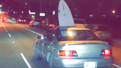 Brown car drives on road with surfboard sticking out of sunroof
