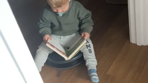 Toddler Reads a Book While Riding Robot Vacuum