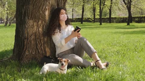 curly young woman in pink face mask sitting on green grass under tree and using mobile phone