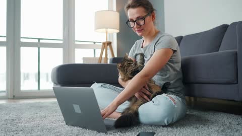 Casually dressed woman sits on a carpet with a laptop, holds on her knees and strokes a fluffy cat