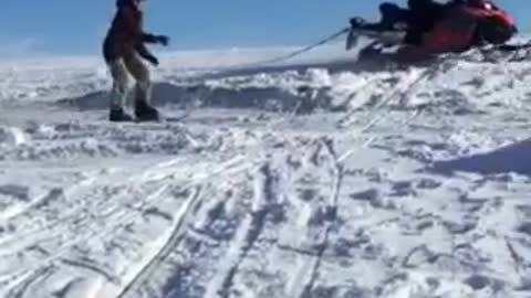 Skier hangs onto rope attached to snowmobile, jumps off ramp and falls