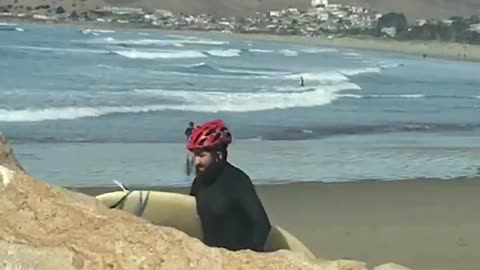 Guy wearing red helmet walking on beach with surfboard