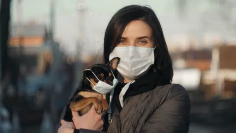 Woman in protective face mask with small dog wearing medical mask too