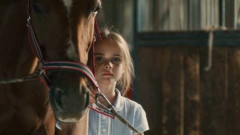 Cute girl holding chestnut horse by bridle and looking at camera