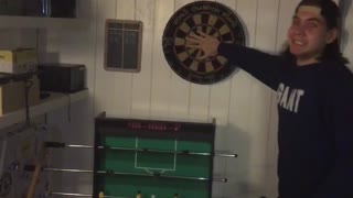 Guy shoots dart in between guy in blue shirts fingers and makes it