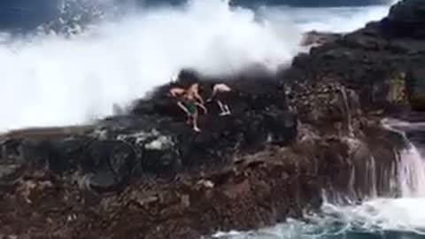 Group of people on top of rocks on beach get pushed into water by waves