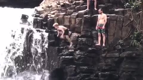Shirtless guy falls off clip next to waterfall bellyflop