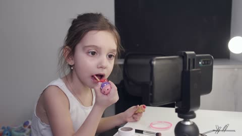 Little Girl Applying Lipstick While Looking At Her Smartphone