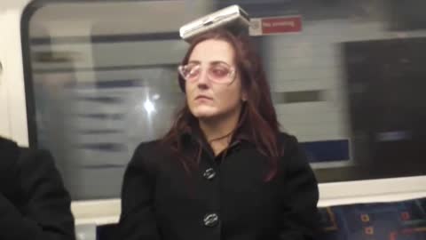 Woman with pink glasses balancing black dog container on her head subway