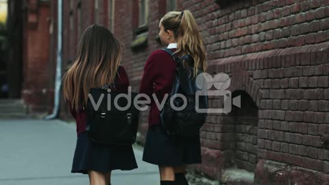 Two Teen Girls In The Uniforms Coming Back From School Home And Walking The Street While Talking