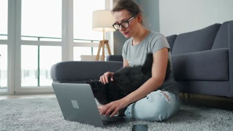 Casually dressed woman with glasses sits on a carpet with a laptop and strokes a fluffy black cat