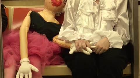 Girl dressed as a mime holds hands with a mannequin doll on subway train