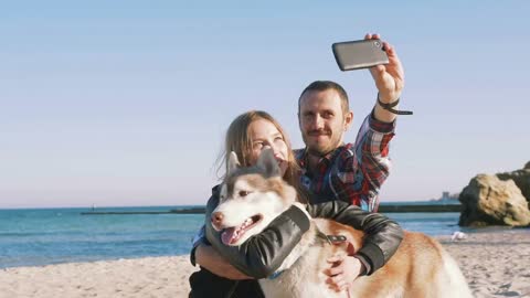 Young caucasian couple on beach doing selfie on smart phone with siberian husky dog