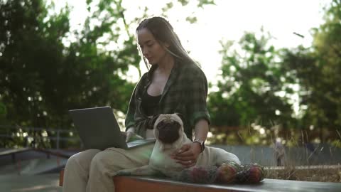 A woman with a laptop sits on parapet in a skate park with her dog pug. Lens flares on background