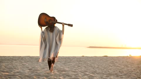 Woman walks on sunset beach with acoustic guitar