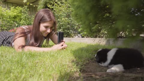 A young beautiful woman lies on grass and takes photos of a cute little puppy under a tree