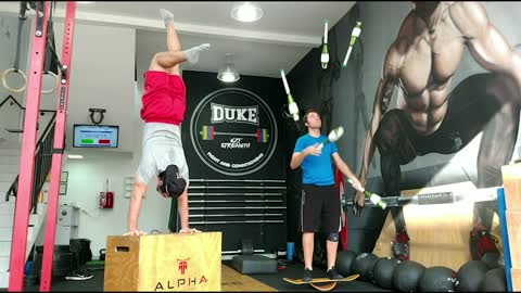Guy Holds A Handstand On A Box While His Friend Juggles Clubs On A Balancing Board