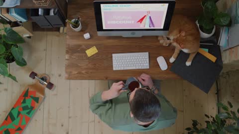 Top-view footage of young man sitting at table with red cat, sipping tea while doing online shopping