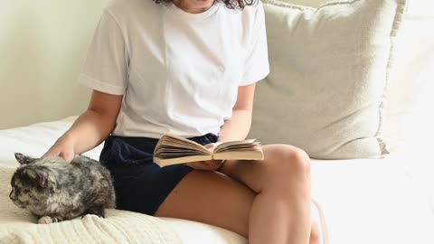 Woman Sitting on the Bed Petting a Cat While Reading A Book