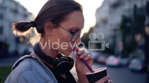 Side View Of A Beautiful Young Woman With Headphones On Her Neck Drinking Beverage Using A Straw