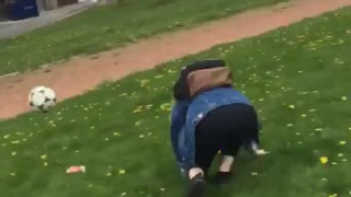 Filmer kicks soccer ball at jean jacket girl and she falls down on grass