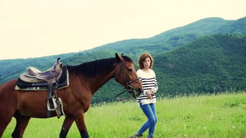 A happy senior woman holding a horse grazing outdoors on a pasture. Slow motion