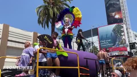 A drag queen standing on a giant eggplant emoji offers lube to a crowd with children in it