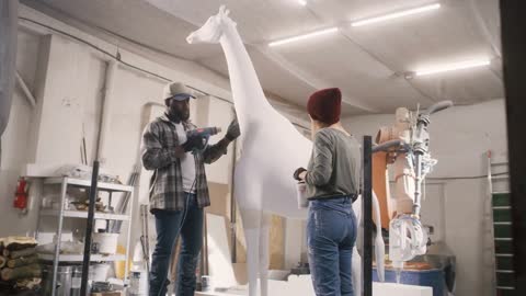 Pan left view of woman mixing paint in cup and checking statue while black man