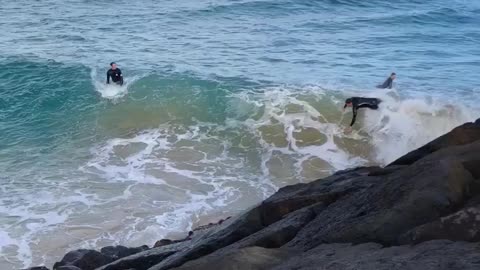 Wet suit guy falls off white surfboard into giant rocks