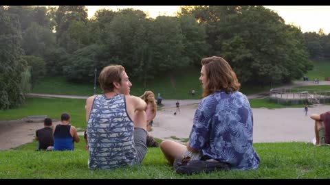 Dogs run around two men having a conversation, while sitting on a hill at sunset