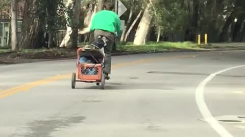 Green guy rides bicycle with wagon attached behind him