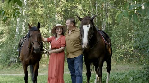 Smiling woman in hat and man with horses in summer forest