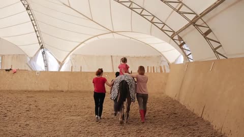 Back view of a little girl learning to ride a horse. An instructor and two women help her