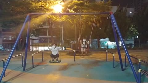 There are two girls riding on a swing in the playground at night.
