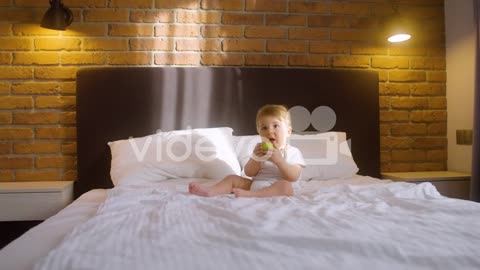 Front View Of A Baby Sitting On The Bed In Bedroom While Bitting A Green Apple